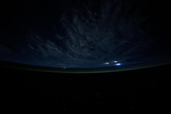 A nighttime view of Earth from space, showing a curved horizon with faint green airglow along the edge of the atmosphere. Above the planet, dark cloud cover is illuminated by isolated bright blue and white flashes of lightning from storms. The black space surrounds the Earth, dotted with distant stars.