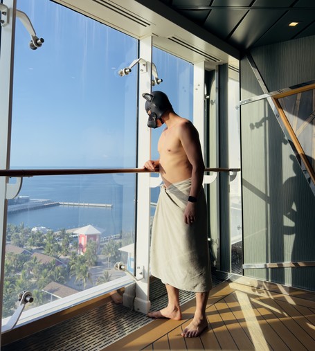 A shirtless person stands barefoot on the polished wooden deck of a cruise ship, wrapped in a towel from the waist down. They lean casually against a glass railing, gazing out through floor-to-ceiling windows at a sunlit coastal port below. The person wears a sleek, black neoprene puppy hood with pointed ears, the mask’s contours catching the warm natural light that streams in from the right. Outside, palm trees cluster around pastel-colored buildings, and a curving pier juts into calm blue water under a clear sky. The scene is bathed in dramatic shadow and golden highlights, creating a sense of relaxed anticipation and playful mystery.