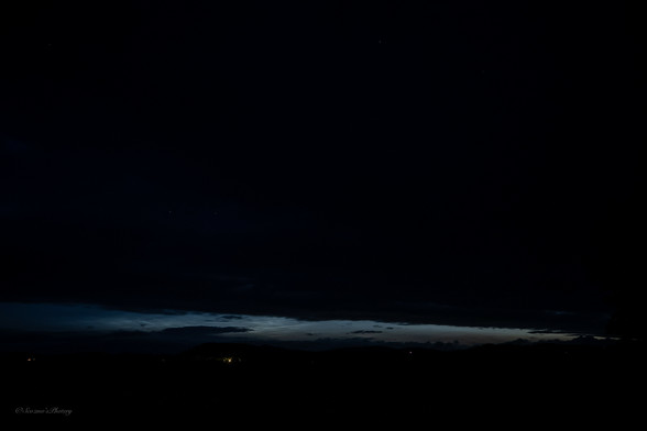 An almost black, night sky image with a pale band of light just above the horizon. Noctilucent (glowing) night clouds can be seen in the pale band. a few scattered house lights can be seen below