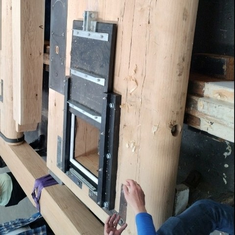 A cropped photo of a pair of oak lock gates in the Bradley Workshop. A pair of hands in the frame shows the massive scale of each oak door—about a foot thick (about 30 centimetres). The door closest to the camera has been shaped, one edge is rounded so the door can rotate once it is placed at one end of the lock chamber.

Added to the lower half of the oak gate is a plastic paddle, about a square yard (about 1 square meter) in size, set in a frame that holds the paddle against a square hole through the oak gate. Essentially, the paddle is a smaller, plastic, sliding door within a massive, rotating, oaken door. But the one door is called a paddle and the other door is called a gate, because historical English just is that way. The tour guide has just slid paddle up to the open position to show how it reveals the square hole through which water will flow to fill (or drain) the lock chamber. The paddle has some steel bracing and a steel bracket for the steel-toothed mechanism and gear (not shown) still to be installed. Boaters will use that mechanism to "wind" or crank the paddle up and down.

The photo is actually rotated to help the viewer imagine the paddle slide up and down when the gate is installed upright. But the gate is actually laying flat on saw horses, and of you look closely at the edges of the photo you'll see people's legs and bellies nonsensically rotated sideways; these are other visitors on the tour of CRT's Bradley Workshop with me.