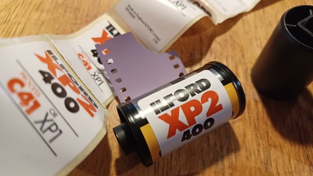 Image of several Ilford XP2 sticker labels for putting on handrolled bulk roll canning and one roll of 35mm film with such a sticker on it.