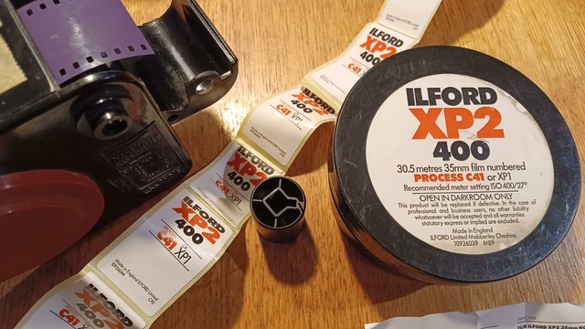 Image of a bulk roller left, several sticker labels for Ilford XP2 film and a plastic circular case that once held 30m of film with the label Ilford XP2 400