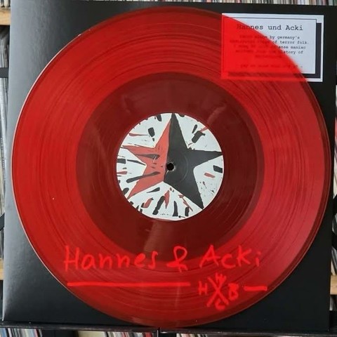 Eine rote Schallplatte mit einem weißen Etikett in der Mitte, auf dem ein Stern abgebildet ist. Die Schallplatte steht auf einem schwarzen Hintergrund, und am unteren Rand steht handschriftlich "Hannes & Acki". Ein kleiner Aufkleber ist in der oberen rechten Ecke.