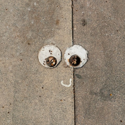 A section of sidewalk where two circular metal pipe fittings filled with debris resemble cartoonish eyes. Below them, a small curved white straw wrapper mimics a smiling mouth, creating a face-like illusion in the pavement.