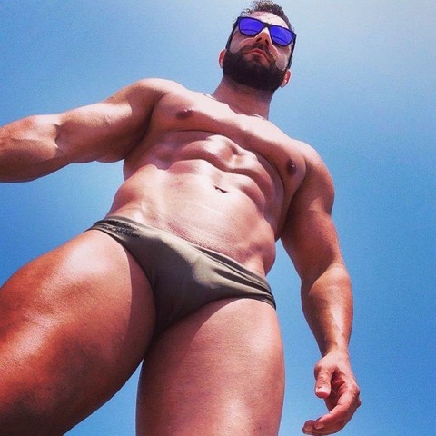 A muscular man wearing sun glasses and speedos is looking down at the camera