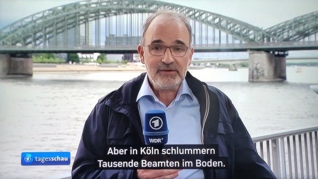 Mann mit blauem Mikro (mit ARD Logo) steht vor Kölner Brücke und dem Rhein. Unten links das Tagesschau Logo. Untertitel-Text: "Aber in Köln schlummern tausende Beamten im Boden."