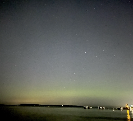 The night sky with a narrow strip of water at the bottom. The lights of a ferry dock on the right side. With stars above there is a horizontal band of green light low in the sky.