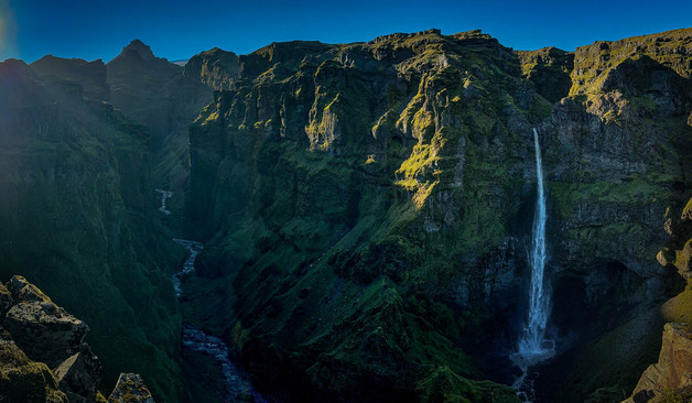 A green mossy canyon with a river running through, a waterfall on the right, and a mountain peak on the left with bright sun coming through the upper left hand corner.