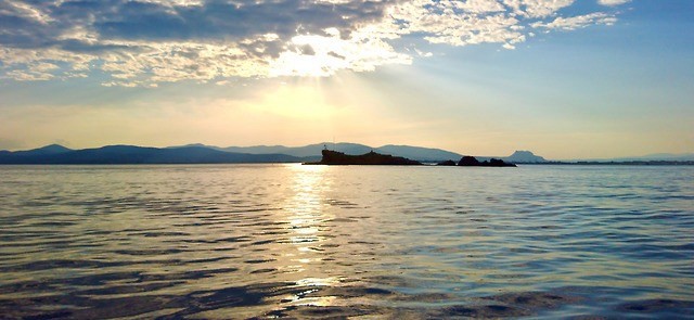 Photo d’un paysage maritime en fin d’après midi. Au premier plan et occupant la moitié basse de l’image : la mer, dont la surface calme reflète le soleil dans une bande blanche bordée d’or, ainsi que les couleurs bleu-jaunâtre du ciel. À l’horizon, un petit îlot de roche et derrière lui, la côte se dessine avec des collines sur la gauche et une plaine sur la droite. 
Occupant la moitié haute de l’image : le ciel, dégradé de bleu allant jusqu’au jaune pâle. Un banc de nuages masque partiellement le soleil de fin de journée, ses rayons se diffusent en étoile au dessus et en dessous des nuages.