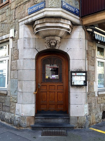 Picture of an arched door, placed on the cut corner of a building. The keystone is ornamented with a lion head.
