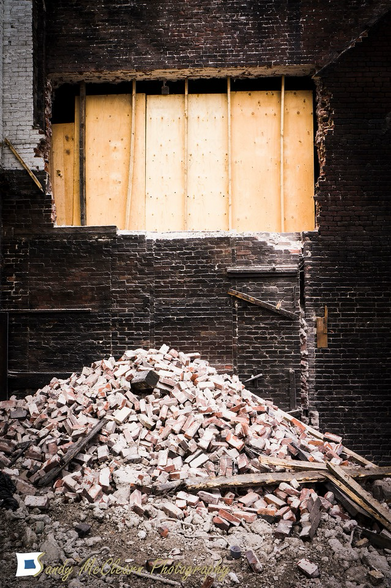 A pile of old bricks and material lies on the ground under an opening broken into the exterior brick wall of an old building.