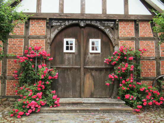 Ein Dielentor aus Holz in der Giebelseite eines Fachwerkhauses. Rechts und links davon üppige Rosenstöcke mit roten Blüten. Das Tor hat zwei kleine, weiße Sprossenfenster. Ein Balkenspruch in dem Balken über dem Tor ist mit weißer Farbe nachgezogen.