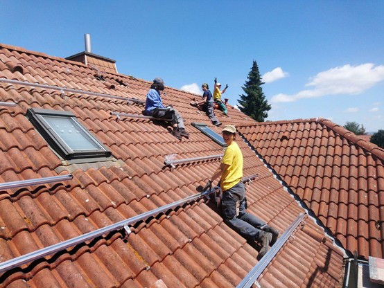 Zu sehen sind mehrere Menschen auf einem Dach, die in die Kamera lachen und dabei Montageschienen für Solarpanele installieren. Der Himmel ist blau und die Sonne scheint.