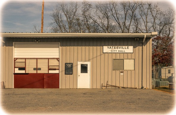 A single-story building with vertical wood siding and a metal roof serves as the "YATESVILLE CITY HALL" as indicated by a sign on the front facade. The building features a large garage door with red trim on the left side, likely housing municipal vehicles or equipment. A white entrance door is positioned in the center, flanked by two small windows. The structure sits on a gravel lot with bare trees visible in the background against an overcast sky. A chain-link fence and utility pole are visible on the right side of the image, giving the scene a small-town, rural municipal appearance.