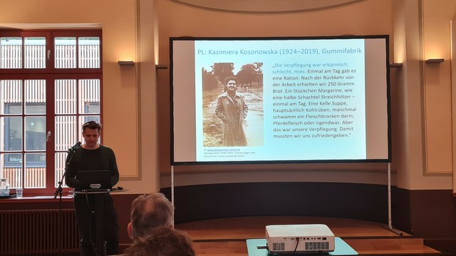 Das Bild zeigt einen Mann, der an einem Laptop steht und eine Präsentation hält. Die Präsentation zeigt ein Foto von Kazimiera Kosonowska (1924–2019), die in einer Gummifabrik arbeitete, und ein Zitat über die erbärmlichen Verpflegungsbedingungen während ihrer Zwangsarbeit. Der Mann scheint ein Vortragender zu sein, der über die Geschichte der Zwangsarbeit in der Zeit von 1939 bis 1945 spricht.  Das Bild ist ein Momentaufnahme einer geschichtlichen Präsentation.