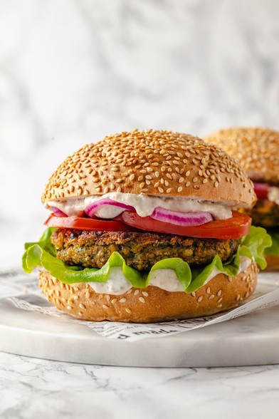Close-up of a vegan falafel burger on a sesame seed bun, layered with lettuce, tomato slices, red onion, and a creamy white sauce, set on parchment paper over a marble surface.