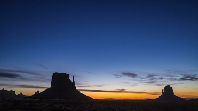 Blue hour at the silhouetted Mittens rock formations in Monument Valley. The sky is deep orange near the horizon while the upper sky is still a dark blue.