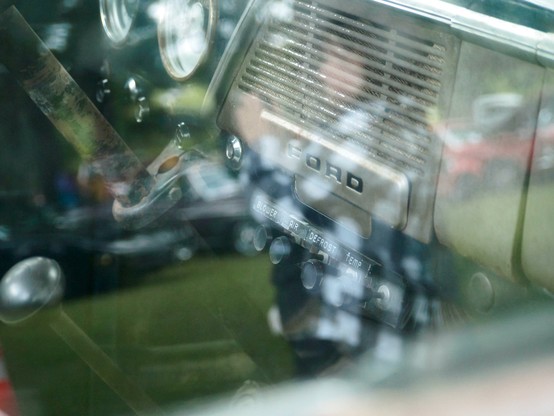 Close-up view of a vintage Ford car dashboard, seen through the car window. Reflections of a person and outdoor scenery are visible on the glass. The dashboard displays classic dials and controls labeled with text, emphasizing the retro design.