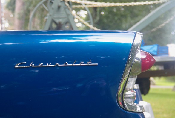 Close-up view of the rear fin of a classic blue Chevrolet car. The focus is on the shiny chrome Chevrolet emblem and the bright red taillight, both prominently featured. The background is outdoors and appears blurred, keeping attention on the detailed features of the cars design.