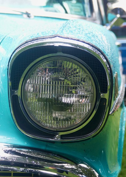 Close-up view of a vintage turquoise car’s round headlight, featuring chrome trim and a portion of the hood. Water droplets are visible on the surface, emphasizing recent rain or washing.