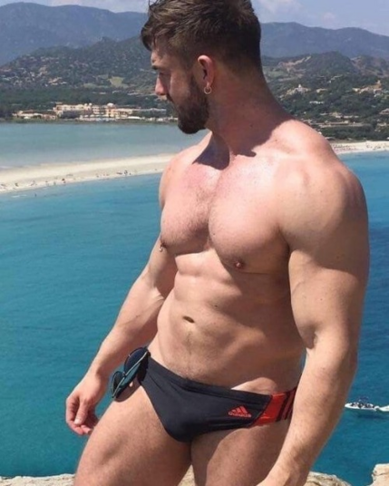 A muscular white man wearing speedos is looking over to the sea from the top of a cliff