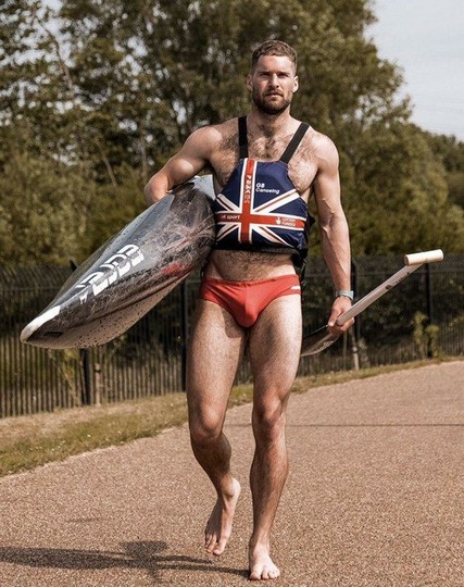 A muscular white man is wearing red speedos and Great Britain canoeing team vest, carrying a canoe
