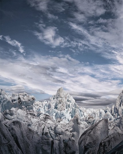 A striking view of a jagged glacier field beneath a dramatic sky. Towering ice formations dominate the scene, their surfaces a blend of frosty blue and weathered grey, suggesting ancient ice shaped by time. The central peak rises sharply like a frozen mountain, surrounded by chaotic ridges and crevices. Above, layered clouds stretch across a blue sky, creating a sense of motion and scale. The overall mood is stark, raw, and awe-inspiring, capturing the harsh beauty of a remote glacial wilderness.