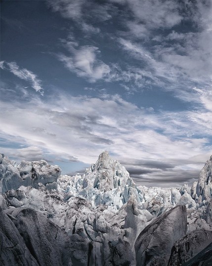 A striking view of a jagged glacier field beneath a dramatic sky. Towering ice formations dominate the scene, their surfaces a blend of frosty blue and weathered grey, suggesting ancient ice shaped by time. The central peak rises sharply like a frozen mountain, surrounded by chaotic ridges and crevices. Above, layered clouds stretch across a blue sky, creating a sense of motion and scale. The overall mood is stark, raw, and awe-inspiring, capturing the harsh beauty of a remote glacial wilderness.