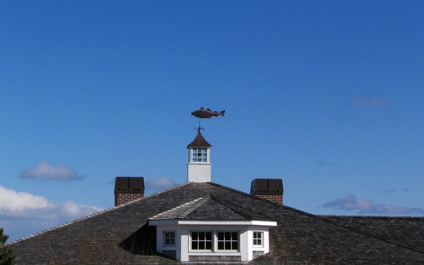 Windows in cupola under a fish-shaped weather vane on the Atlantic coast of Massachusetts, wooden-shingled roof and brick chimneys against blue sky with some white puffy clouds.