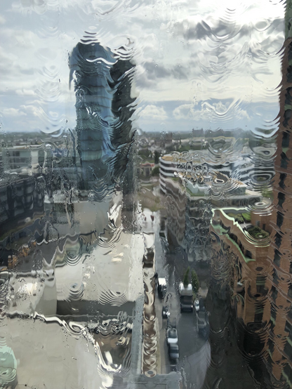 Blick durch an der Fensterscheibe herunterrinnendem Regenwssers auf die Gebäude im Medienhafen Ddorf. Rechts die rote Fassade eines Wohnhochhauses, mitte rechts ein rundes und eckiges Geschäftsgebäude, mittig links die schuppige, bläuliche Glasfassade eines Hochhauses. Durch das Regenwasser auf der Fensterscheibe sind die Gebäude zum Teil stark verzerrt.