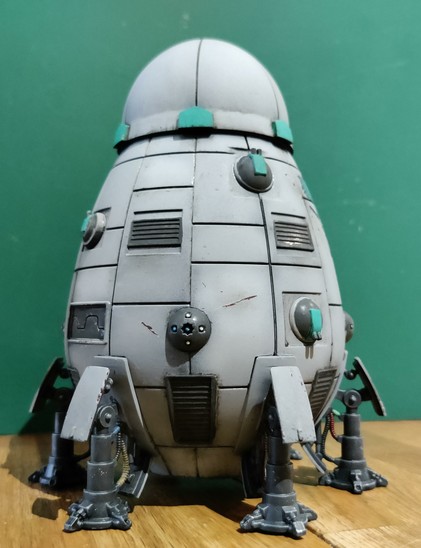 A low angle photo of the grey Overlord-C DropShip on a wooden table and a green cutting mat's backside as a background. No faction markings are visible. The grey hull has some random scorchmarks from lasers and impact marks from autocannon hits painted on.