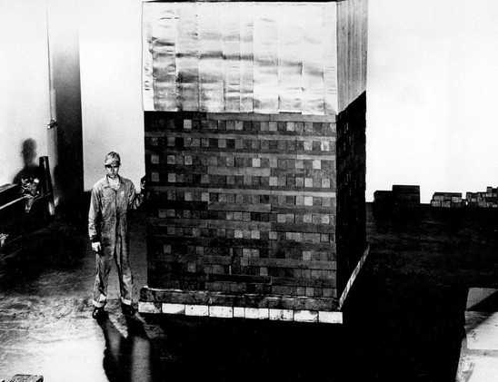 Ein Mann in einem overallartigen Arbeitsanzug steht neben einer großen, rechteckigen Struktur, die aus gestapelten, rechteckigen Blöcken besteht. Die Struktur scheint aus verschiedenen Materialien zu bestehen, und der Mann hat seinen rechten Arm an die Seite der Struktur gelegt. Die Umgebung ist schlicht und leer, auf dem Boden sind reflektierende Flächen zu sehen. Die gesamte Szene ist in Schwarzweiß gehalten.