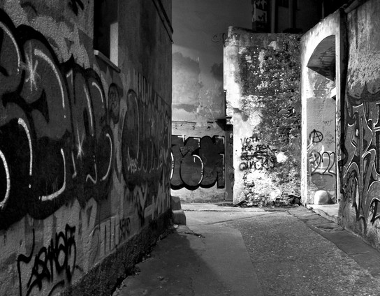 The image depicts a narrow alleyway at night, captured in black and white. The walls on either side are covered in graffiti, with various tags and designs in different styles and sizes. The left wall features large, bold graffiti with intricate designs, while the right wall has more scattered and smaller tags. The alley is dimly lit, with shadows cast by the surrounding buildings, creating a moody atmosphere. The ground is a concrete path, and the walls show signs of wear and age, with peeling paint and exposed bricks. The background reveals more graffiti-covered walls and an arched doorway, adding to the urban feel of the scene.

Provided by @altbot, generated privately and locally using Ovis2-8B

🌱 Energy used: 0.153 Wh