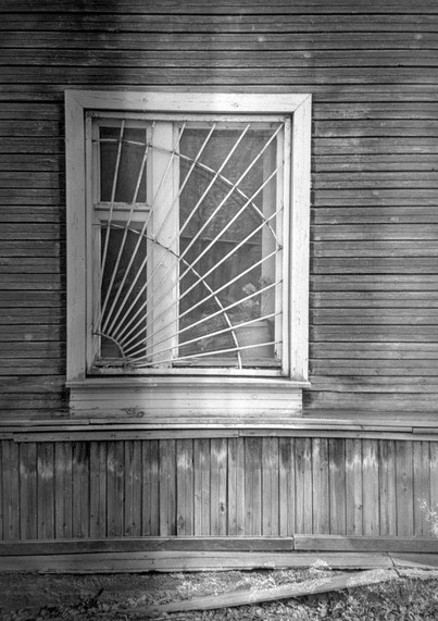 Monochrome photo shows a close-up of a window of an old wooden house. There is a metal grate in the form of a spider's web on the window.