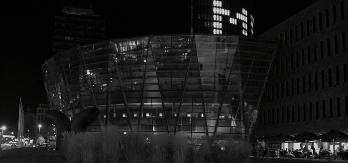 A modern glass building with curved architecture shines in the city at night, while people sit outside a nearby restaurant, surrounded by the urban darkness.

Ein modernes Glasgebaeude mit geschwungener Architektur erstrahlt nachts in der Stadt, waehrend die Menschen vor einem nahe gelegenen Restaurant sitzen, umgeben von der staedtischen Dunkelheit.