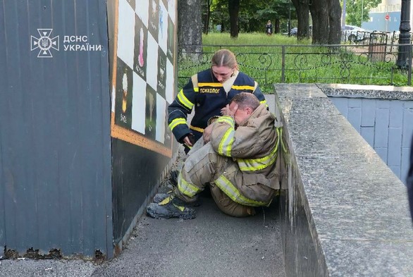 A woman comforts a man prostate in grief in an alley, both wearing rescue uniforms