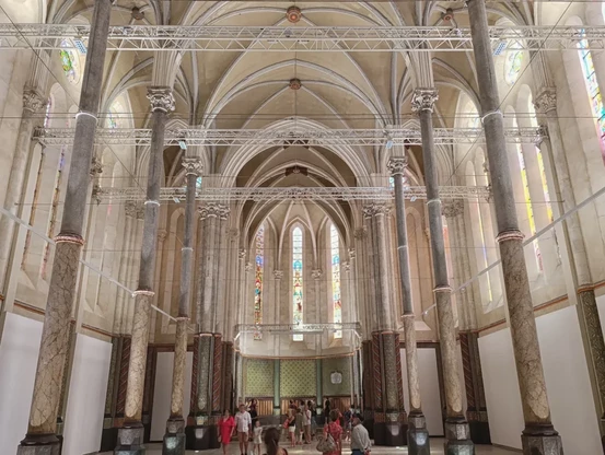 Photo de l'intérieur d’une église reconvertie en lieu d'exposition, avec de grandes voûtes en ogive, des piliers en pierre, des vitraux en hauteur et une structure métallique suspendue. Au bas de l'image des visiteurs.