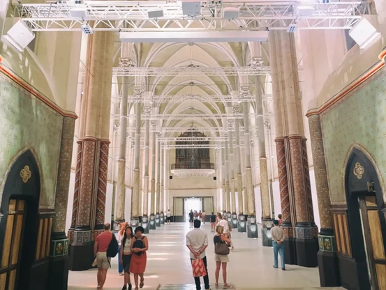 Photo de l'intérieur d’une église reconvertie en lieu d'exposition. La nef centrale est encadrée de piliers décorés, les murs sont clairs avec des motifs peints, et des personnes se déplacent dans le couloir. Au fond, on perçoit l'ancien orgue au-dessus de l'entrée.