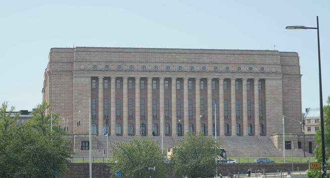 A photograph of a tall, light-colored stone building featuring 14 columns on its front. A wide set of steps lead to them. It is the parliament house of Finland in Helsinki. The picture was taken from afar, so the building is in the background, being car traffic, streets lights, and tree tops.