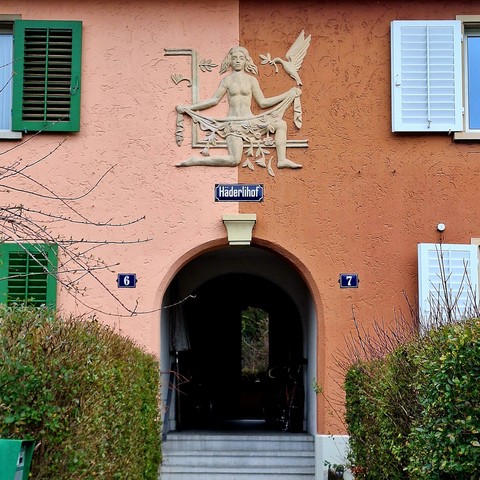 Picture of a passageway between two housing units. Above, a sign reading "Häderlihof" and the bas-relief of a nude woman with flowers.