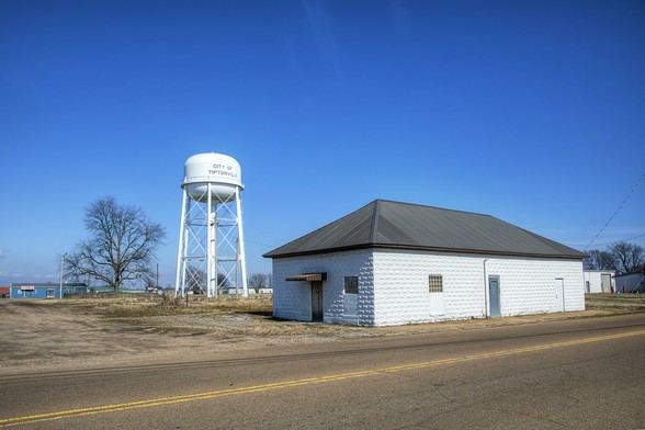 A white water tower marked "City of Tiptonville" rises above a flat landscape near a small building with a metal roof.
