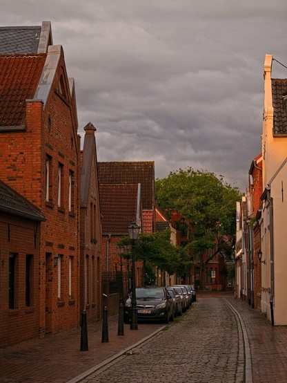A slightly curved road. It consists of cobblestones and is narrow. A few cars are parked in a row on the left-hand side. To the left and right are single-storey houses, red brick on the left and painted a light colour on the right. Warm sunlight falls on the houses on the right. The sky above the houses is overcast with grey clouds. At the end of the street is a large, green tree.