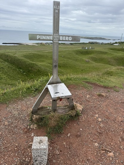 A wooden cross marking the height of 61.3 meters, labeled "PINNEBERG," is situated on a hilltop with a view of the sea. The landscape features green hills and a coastal area in the background.