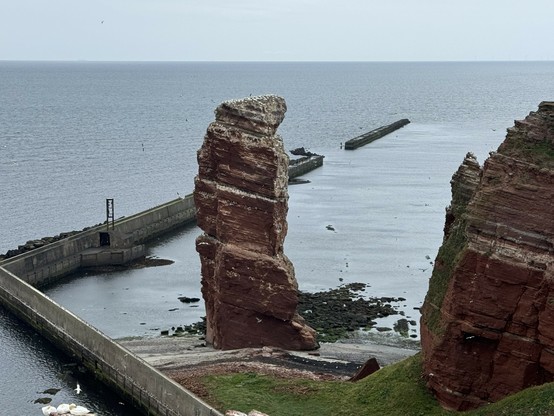 A tall, red rock formation stands prominently by the sea, surrounded by calm water. In the background, a harbor wall extends into the ocean, and there are some scattered rocks visible. The sky is overcast, adding a serene atmosphere to the coastal view