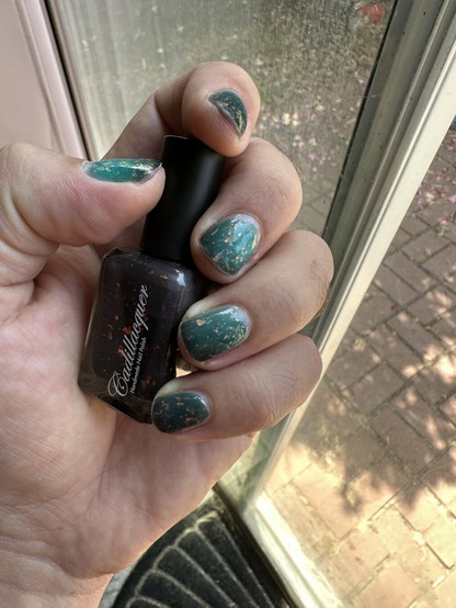 A hand holds a bottle of nail polish next to manicured nails featuring a teal color with gold flakies and a gradient. The background shows a window and brick pavement outside.