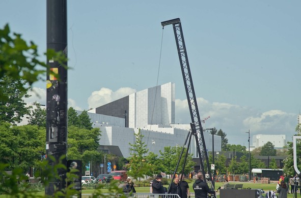 A multi-layered photograph of the eastern side of Finlandia Hall, an angular and white building. On the foreground out-of-focus foliage, on the mid-ground a small crane operated by a crew of workers preparing a stage and passers by.

The photo makes it seem like the photographer was hiding when the shot was taken.
