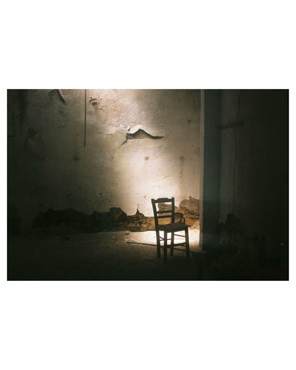A dark abandoned room with a small sunspot on the floor and reflecting on the wall. The room is dusty and empty except for one chair standing in silhouette in front of the light spot. The plastering is flaking off the wall; at ground level you can already see the original stone wall.