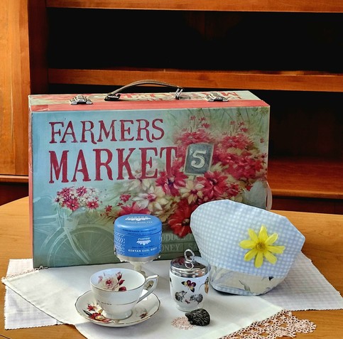 Tea tableau in hazy afternoon colors in front of wooden shelving, with a cardboard suitcase with an old- timey design of Farmers Market flowers in a bicycle basket. It rests on a gingham sheet and a lace-trimmed tea towel, with a gingham tea cozy, rose- patterned vintage china, a jam jar with a garden scene with hedgehogs, next to a hedgehog miniature figurine, and a small pedestal holding Kenyan Earl Grey loose-leaf black tea.