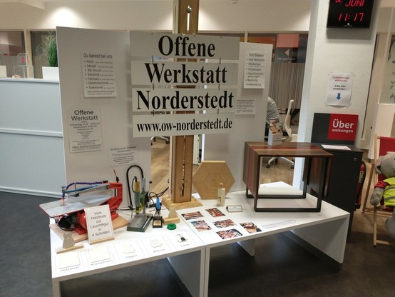 Auf einem weißen Tisch sind Werkzeuge (Dekupiersäge, kleiner Bohrfräser im Bohrständer und Lötkolben) sowie diverse Werkstücke aus Holz (u.a. ein Tisch) und Fotos ausgestellt. Im Hintergrund werden die Themenbereiche und Möglichkeiten der Offenen Werkstatt Norderstedt genannt / beworben.