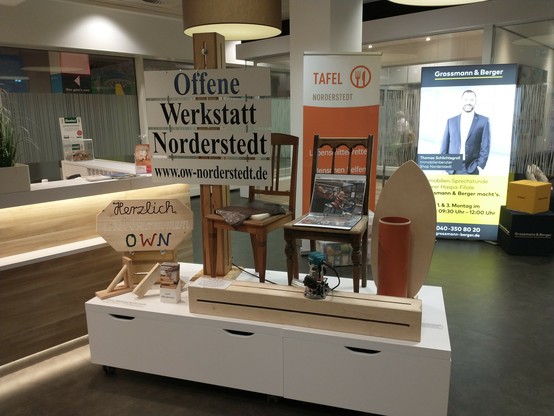 Auf einem weißen Podest mit Schubladen ist Werkzeug (Oberfräse) sowie diverse Werkstücke aus Holz (u.a. ein zu reparierender Stuhl und ein Brett in welches mit Hilfe der Oberfräse Schrift eingefräst worden ist) und Fotos ausgestellt (Ordner mit Bildern aus unserem Bereich Fahrrad). Im Hintergrund wird die  Offene Werkstatt Norderstedt genannt / beworben.